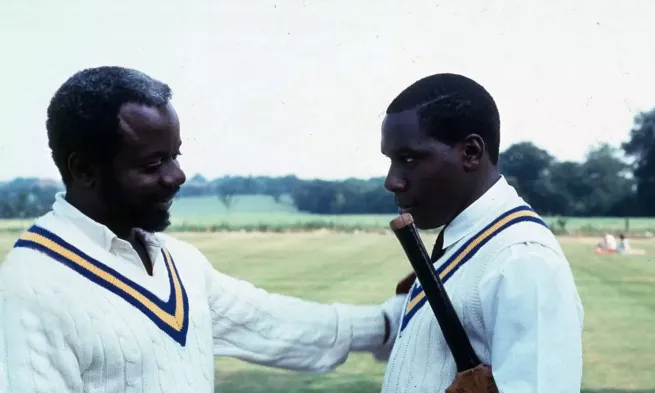 An older man faces a younger one and looks at him fondly. They are both wearing cricket clothes and are on a playing field.