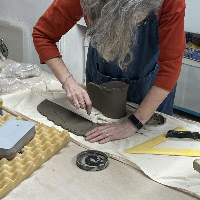 A person leaning over a slab of clay, scoring it with a tool.