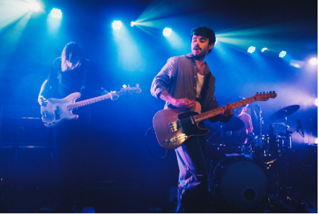 The image shows Cian Godfrey performing live with Somebody’s Child. Cian is at the front of the stage, preforming passionately playing guitar. Behind him, the band members are lit by atmospheric stage lights, with drums visible. 
