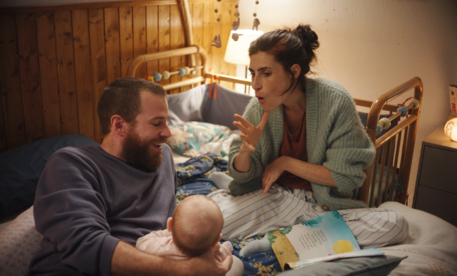 A man and a woman sit on a bed with a baby. The woman faces towards the camera and is shown speaking with sign language. 