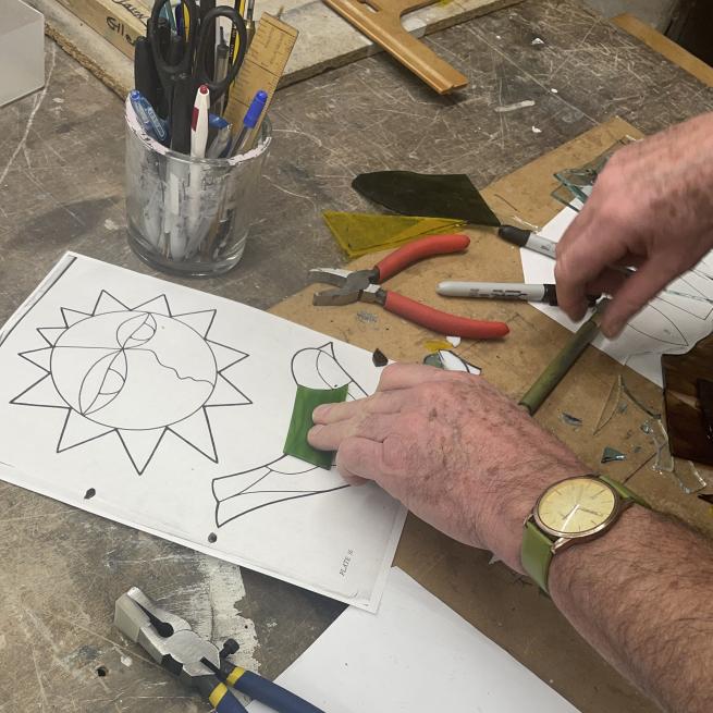 a paper template of a sun and bird with two hands holding a piece of green glass in place on the bird