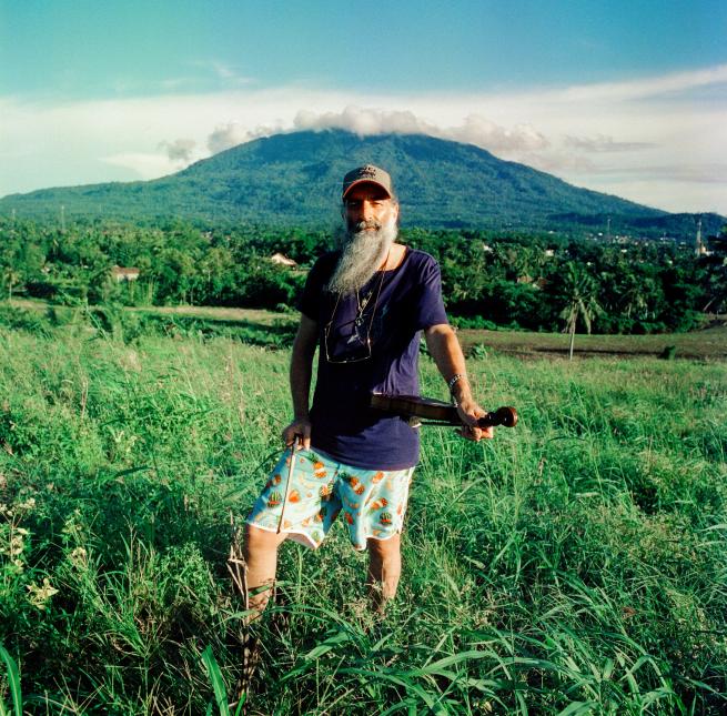 A man with a long beard stands in an open field. Pictured behind him is a mountain. 