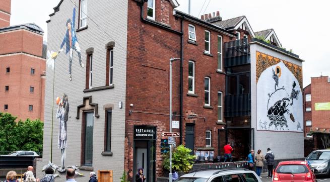 An image of a large building at East Street Arts, Convention House, with two murals visible in the image. To Be or Not To Be, a mural by Taqi Sateen on the left gable wall, and Theft on the last Monday of the year by Mohammad Barrangi on the right wall. 
