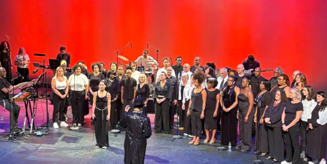 A choir and orchestra perform on stage, illuminated by dramatic red lights.