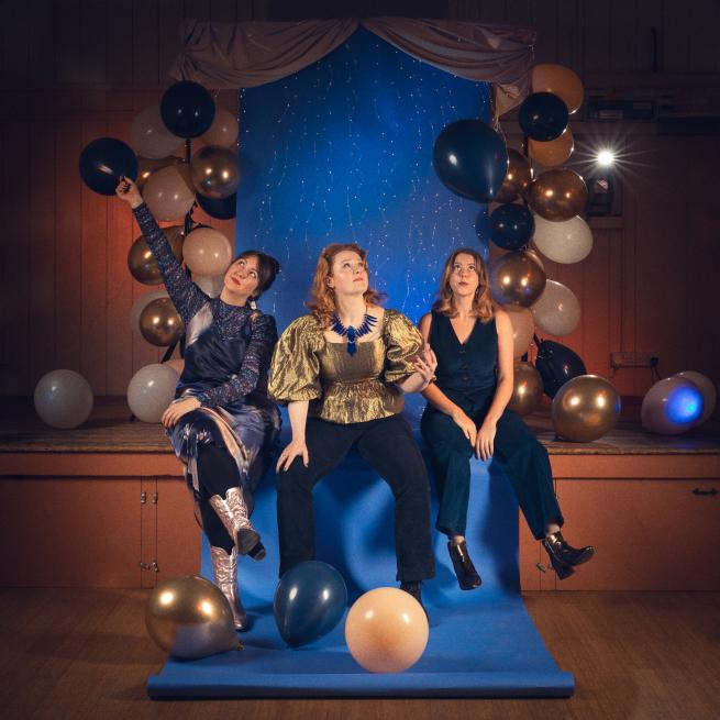 3 women sit on a village hall stage surrounded by gold, white and balloons.  They wear sparkling gold, blue and purple outfits and look up to the ceiling.