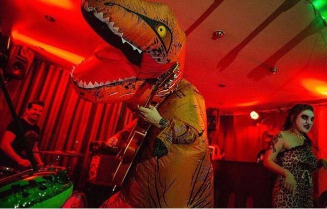 Inflatable dinosaur plays guitar on a red lit stage.