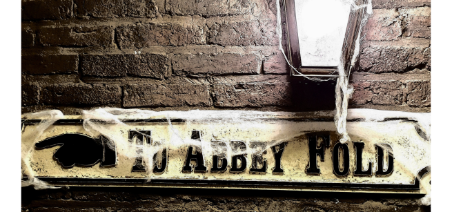 A sign post which says ''To Abbey Fold' covered in spooky cobwebs, on a brick wall next to a Victorian street light