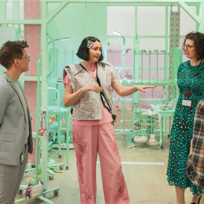 A person with a short black bob wearing a green patterned vest over a pink top, stood in a pale green installation talking to a group of people.