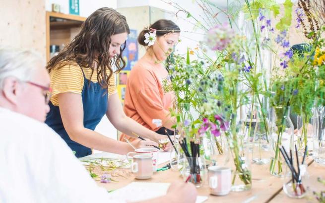 Three people sat or stood around a table drawing flowers in glass vases.