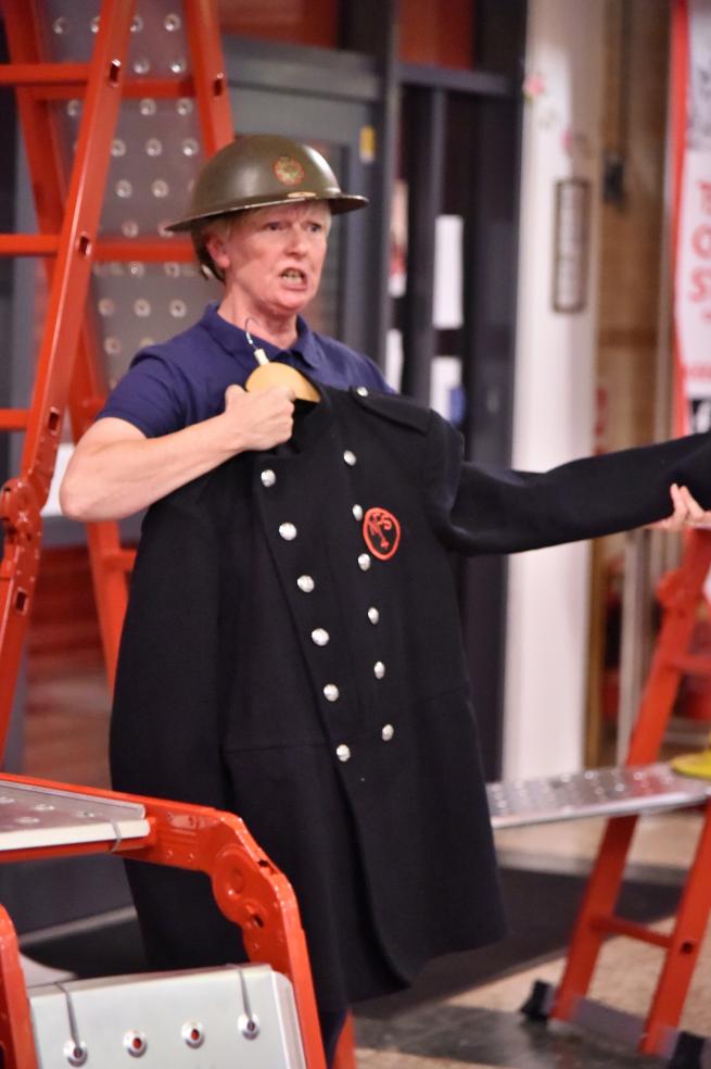 Performer holding a WW2 fire uniform up to themselves with a WW2 helmet on 
