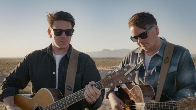 Two brothers with dark hair wear sunglasses and hold acoutic guitars. An American desert landscape is in the background. 
