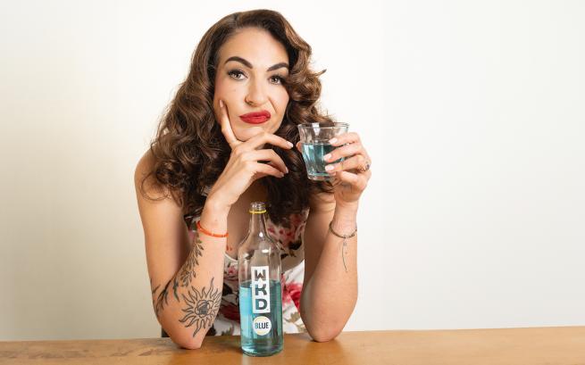 British-Lebanese comedian Esther Manito's long brown hair covers her shoulders, her elbows rest on the table displaying tattoos on her arm, as she looks straight to camera looking really fed up and holding a blue alcopop.