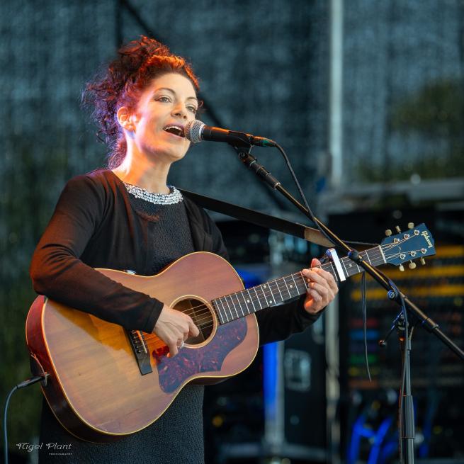 Edwina, a woman with dark hair, stands singing in front of a microphone and playing an acoustic guitar. 