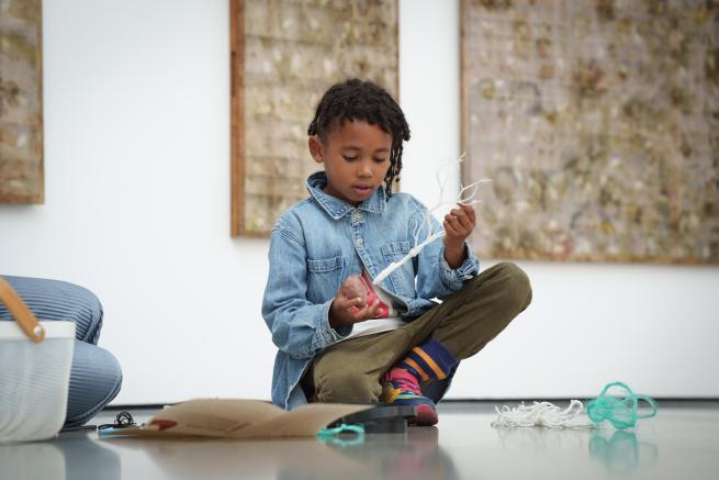 A child sat on the floor of an art gallery, playing with a white twig