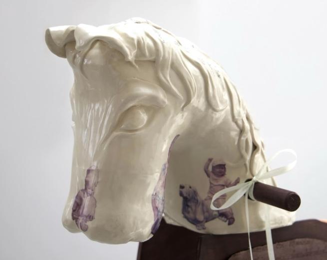 Image depicts a large ceramic rocking horse head with wooden handles. On the horses nose, there is a small image of a baby doll, and on the neck there is a small image of smiling child with a dog. There are white ribbons tied around the wooden handle. 