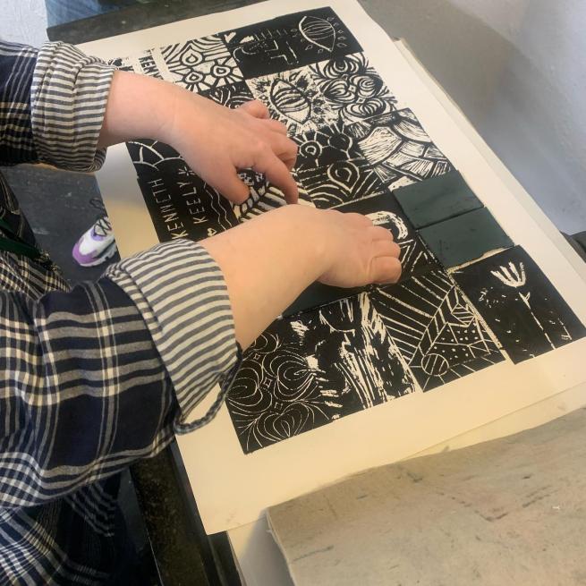 Two hands holding down a piece of lino while working on a large black and white print.