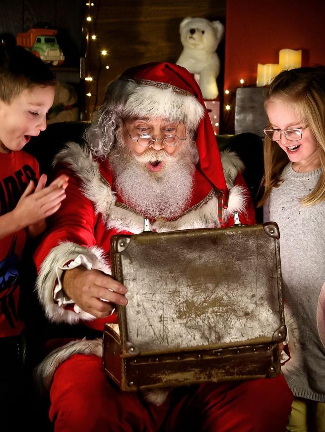 Santa Claus sits with two children looking into a case. They seem excited at what is inside.