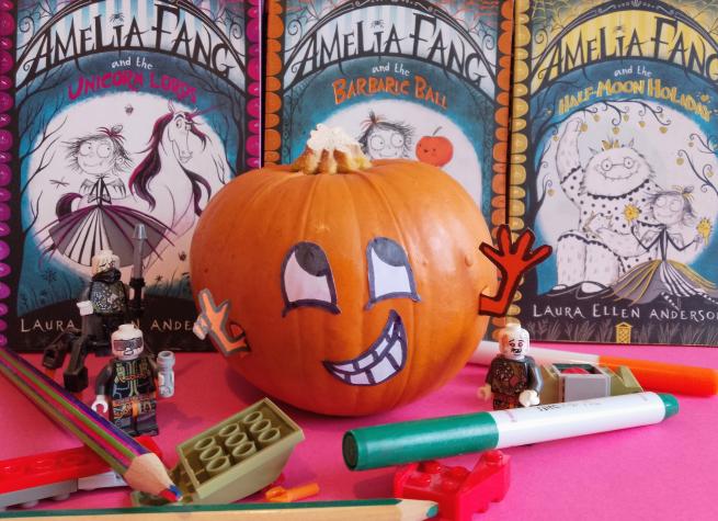 A pumpkin with eyes, arms and mouth is surrounded by craft materials and books