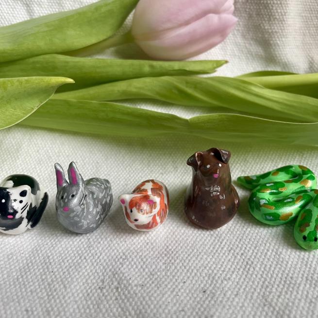 5 polymer clay animals sit on a beige cloth with a pink tulip behind them