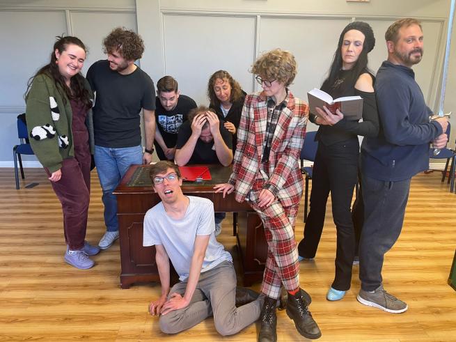 A group is gathered around a desk.  To the right a woman stands reading a book.  Her back is against a man holding a sword.  A man and a woman stand on the opposite side of the desk close and smiling at each other.  There is a man on the floor looking scared and another man with his head in his hands sat at the desk.  Behind him, three other people look concerned for him.