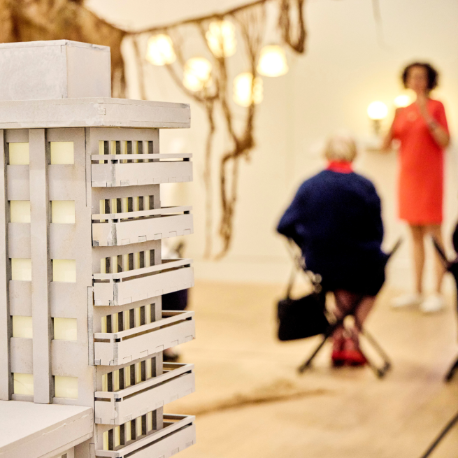 Close up of an architectural model of a block of flats. Behind it there are lights hanging from the ceiling, a large hessian sculpture, and a group of people listening to a talk.