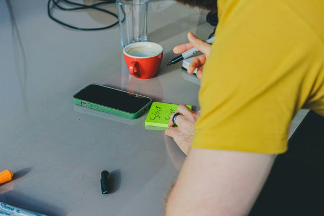 Photo of someone writing on a yellow post-it note at a table, with a coffee cup and mobile phone nearby.