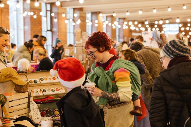 Person looking at embroidered items on a Christmas market stall. A young person wearing a red and white Santa hat has their back to the camera. 
