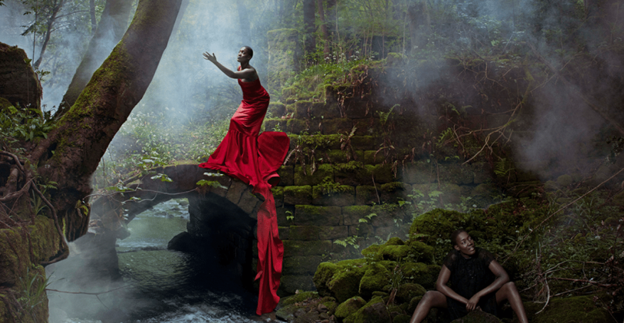 A woman in a wood wearing a red dress stood on a rock, the same woman wearing black sat on the mossy ground