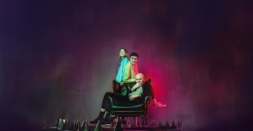 Three people sitting in, leaning on and standing behind a leather chair surrounded by what looks like a metal trap. 