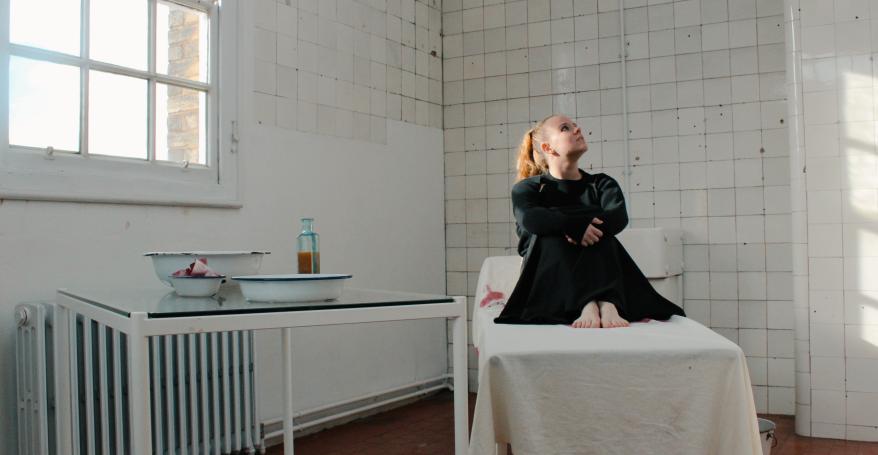 Playwright and musician Amanda Grace sitting on a bed in a tiled Victorian hospital.