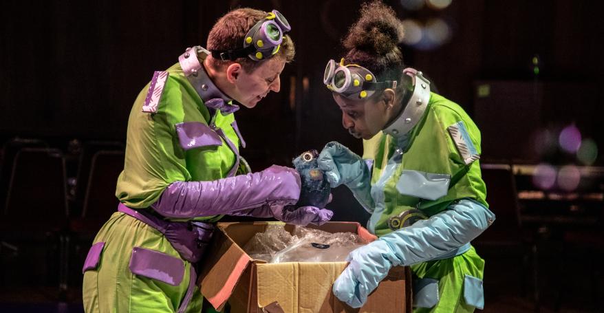 Two people wearing green and purple jumpsuits, looking intently into a cardboard box