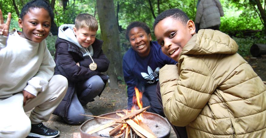 Three children in the woods, sitting round a campfire, smiling at the camera.