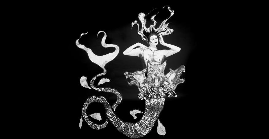 A monochromatic mermaid stands against a plain black backdrop. Her eyes are closed, hands framing her face and has long flowing hair. Her tail is long and spindly with three snarling dog heads placed at her waist.