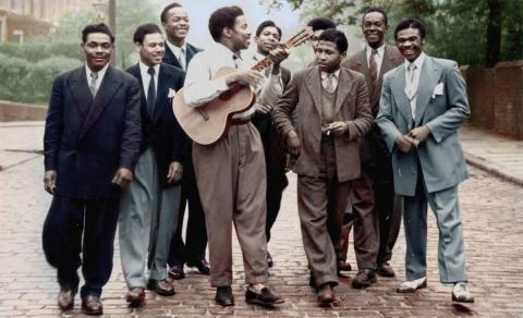 nine men walking, one holding a guitar