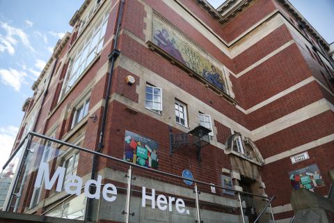 Looking up at red brick bulding with glass barrier outside with the words Made Here printed on. 