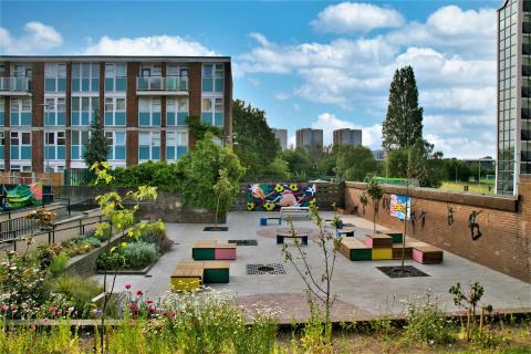 A photo of the Roxby Community Garden. A green community space with colourful seating, surrounded by grassy banks and lush flower beds in the heart of Lincoln Green.