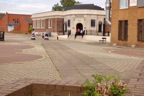 In there foreground there is a wide open town square, leading towards a building. The building is single-storey and is made of pale stone. It has stairs and a ramp leading up to the entrance. On the front of the building is a sign which says Hunslet Community Hub and Library. A mum with two children is exiting the building through the front doors. Two more mums are walking towards the camera, each with a baby in a pram and an older child walking next to them. 