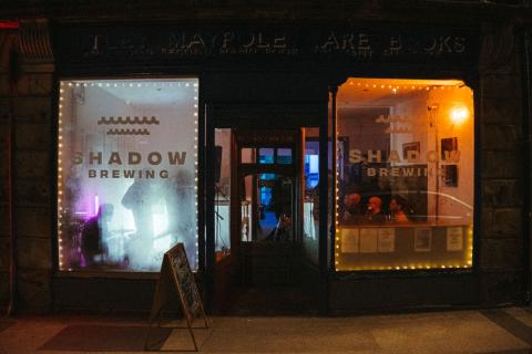 The two shopfront windows of Shadow Brewing are illuminated in an otherwise quite dark photo. In the left had side window, a band is playing, while in the right, an audience sits and watches.