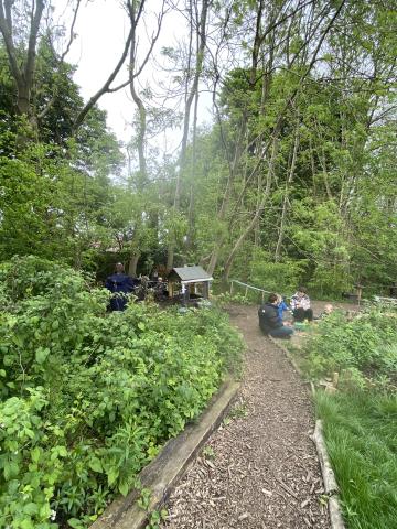 A garden path leads to a natural sandpit, a children’s play house and mud kitchen. Planting is green and wild. 2 mums sit chatting whilst a toddler plays in the sand