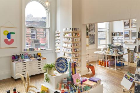 A photograph of the shop which is filled with brightly coloured homeware and cards, bright light is shining in the white painted space through the beautiful arched sash Victorian windows.