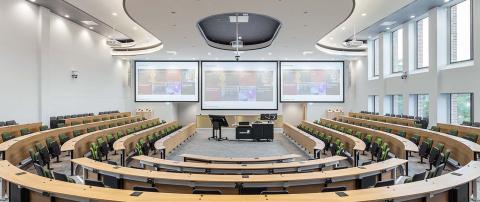 A modern lecture hall with tiered seating, two large projection screens, a lectern, and windows along one side.