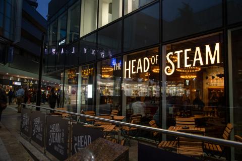 An exterior picture of the Head of Steam Bar on Park Row, Leeds. With large glass windows and the Name in illuminated letters on the outside.