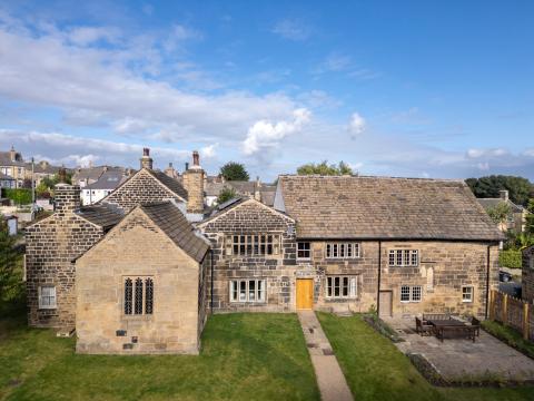  An aerial shot shows Calverley Old Hall, a historic stone building with multiple sections and varying rooflines, under a partly cloudy sky. In front of the hall is a well-maintained green lawn with a light-colored gravel path leading to a wooden front door. To the right of the path, a stone patio with a large wooden table and benches is visible. In the background, other stone buildings with rooftops are nestled among trees, suggesting a village setting.