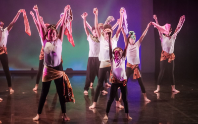 A group of young dancers of mixed ethnicity holding bandanas facing the front with their arms up and smiling.