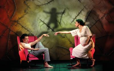 A man and a woman sit in red arm chairs. They reach towards each other extending their index fingers.