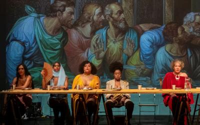 A reinterpretation of ‘The Last Supper’ painting with people gathered around a table. In the background, the original artwork. 