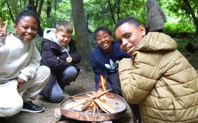 Three children in the woods, sitting round a campfire, smiling at the camera.