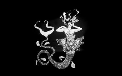 A monochromatic mermaid stands against a plain black backdrop. Her eyes are closed, hands framing her face and has long flowing hair. Her tail is long and spindly with three snarling dog heads placed at her waist.