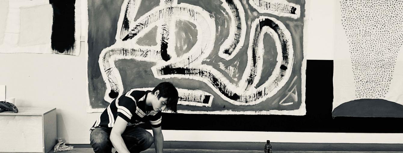 A person paints on a work on the ground with an abstract painting hung behind them