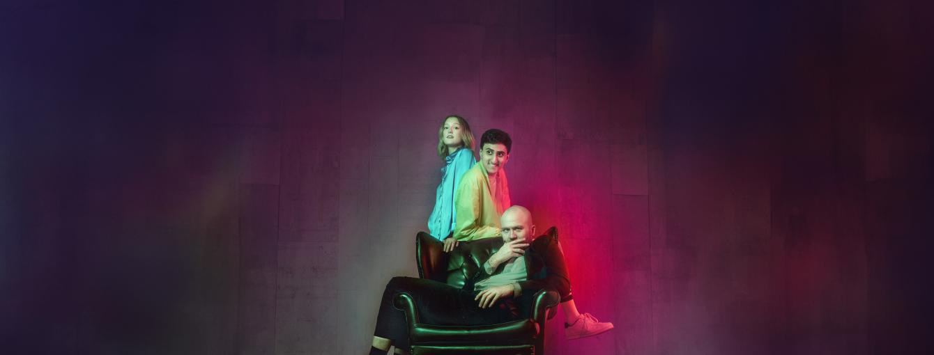 Three people sitting in, leaning on and standing behind a leather chair surrounded by what looks like a metal trap. 
